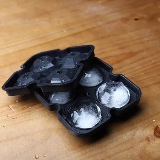 Black silicone ice cube mold open showing four large faceted spherical ice cubes with small circular holes in the center, mold resting on wooden surface with water droplets