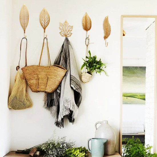Set of five golden metal leaf-shaped wall hooks with different leaf designs holding bags, a blanket, and a plant