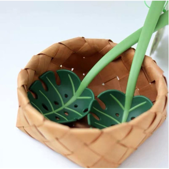 Green Monstera Leaf Plastic Serving Spoons Two green plastic serving spoons shaped like monstera leaves with holes in a woven basket