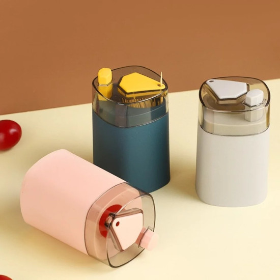 Three automatic toothpick dispensers in pink blue and white with transparent tops Three automatic toothpick dispensers in pink, blue, and white colors with transparent tops. Each dispenser has a triangular button and a small hexagonal knob on the top. The blue dispenser has a toothpick standing upright near the hexagonal knob. The pink