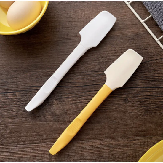Two long silicone spatulas with flat heads, one white and one yellow, on a wooden surface