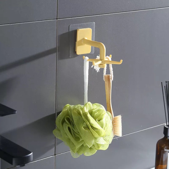 Yellow rotating hook mounted on a dark tiled wall with four short arms extending horizontally. Two items hang from the arms: a green bath sponge with a white loop and a small wooden brush with a white rope loop. The hook is attached to a transparent adhes